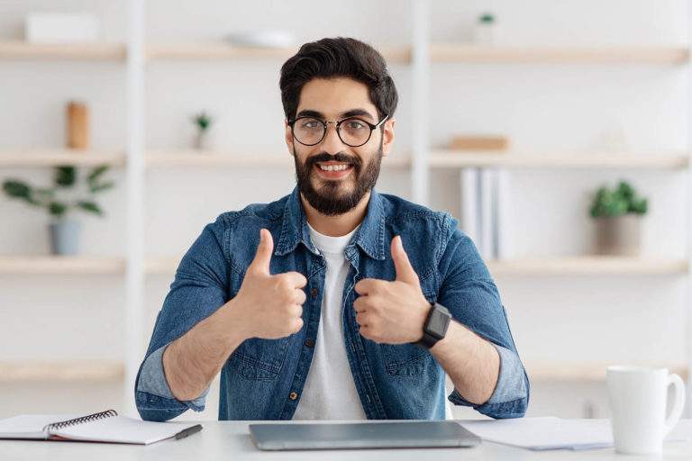 Distant job opportunities. Excited arab guy showing thumbs up gesture while sitting on workplace with laptop. Eastern man enjoying remote online work and smiling to camera